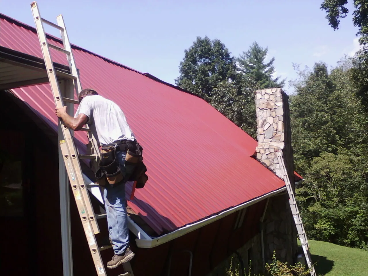 Roofer on a ladder beside a red metal roof and stone chimney — Soffit & Fascia Repair by Roofing Brothers in Darby